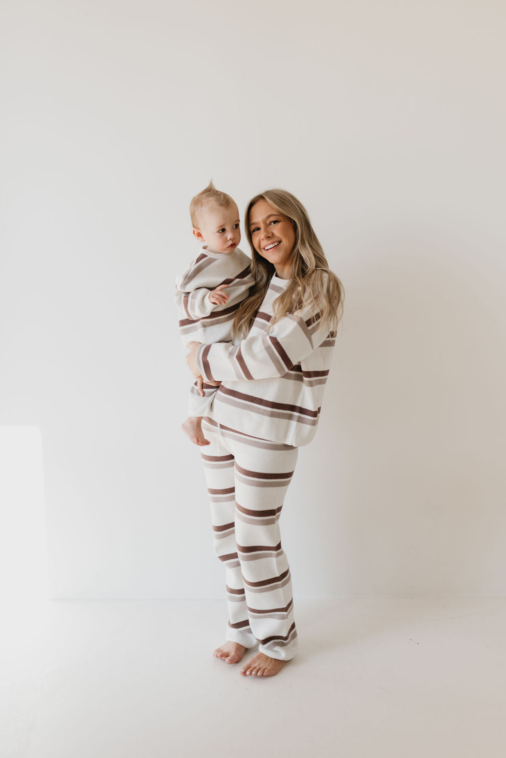 A woman and a baby stand against a white background, both wearing the "Knit Pant Set | Woodland Stripe" from forever french baby, perfect for a cozy fall wardrobe. The woman smiles while holding the baby, who gazes curiously to the side. These matching outfits are crafted from 100% cotton for ultimate comfort.