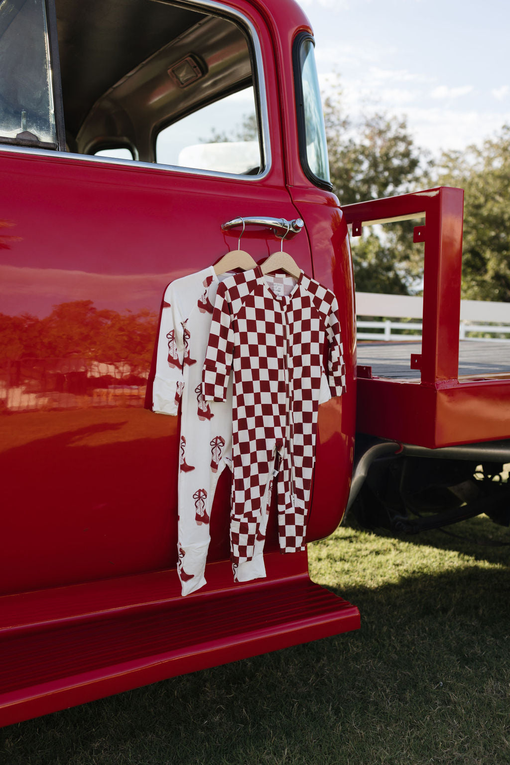 Hanging on a red vintage truck are two adorable garments: one is white adorned with red dog prints, and the other features a maroon and white checkered pattern. Crafted from hypo-allergenic fabric, these Bamboo Zip Pajamas from lolo webb's Cowgirl Christmas collection are perfect for sensitive skin. The outdoor setting includes grass and a fence in the background.