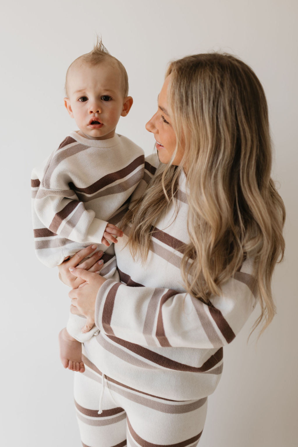 A woman holding a baby, both dressed in matching Women's Knit Pant Set | Woodland Stripe from forever french baby, exude warmth against the plain white background. Her long blonde hair complements the baby's little mohawk. This cozy scene perfectly captures the essence of a fall wardrobe crafted from 100% cotton.