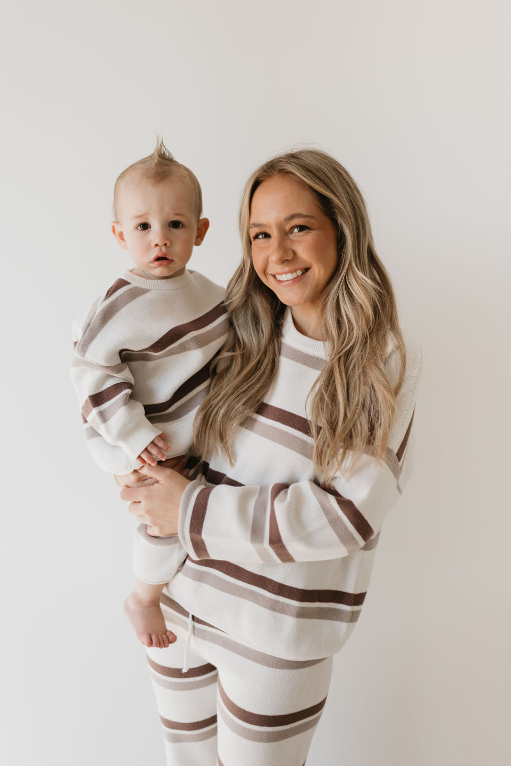 A woman holding a baby, both wearing the Women's Knit Pant Set in Woodland Stripe by forever french baby, stand against a plain white background. Made from 100% cotton, these outfits are perfect additions to your fall wardrobe as they smile warmly at the camera.