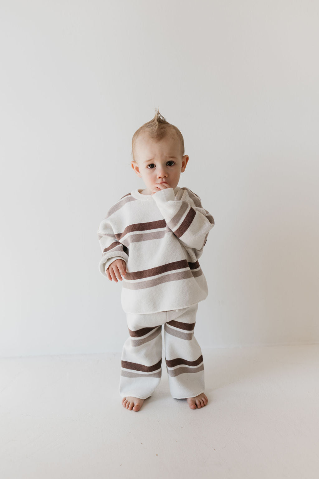 A baby stands barefoot on a white surface, wearing the Knit Pant Set in Woodland Stripe from forever french baby. The cozy cream and brown stripes, crafted from 100% cotton, complement the child's thoughtful expression and small tuft of hair sticking up against a plain, light background.