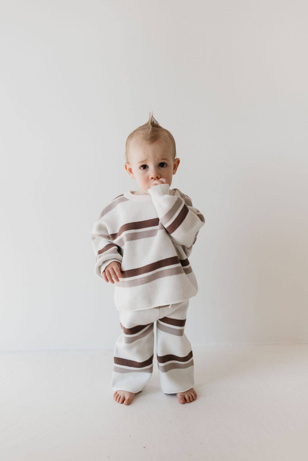 A toddler with a small mohawk stands barefoot against a plain white background, wearing the Knit Pant Set in Woodland Stripe by forever french baby. The cream outfit features brown horizontal stripes and is made from cozy 100% cotton, making it perfect for a fall wardrobe. The child holds one hand near their mouth, creating a shy or thoughtful expression.