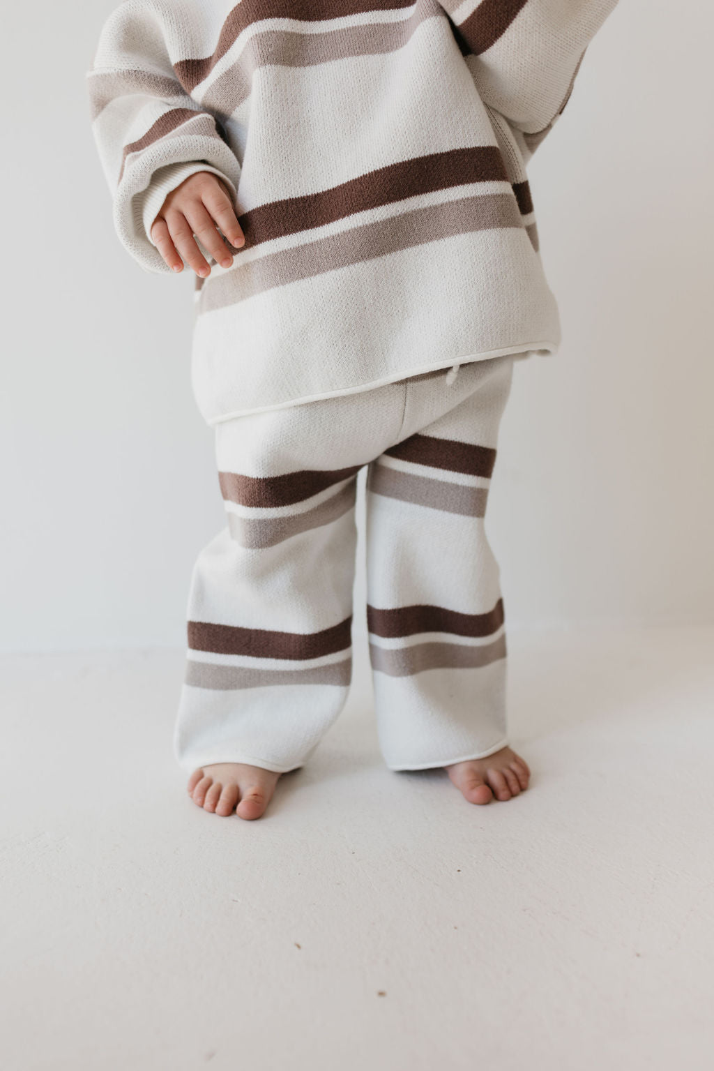 A small child stands barefoot on a light-colored floor, wearing the Knit Pant Set in Woodland Stripe from forever french baby. The cozy ensemble, with its wide brown and beige stripes, is perfect for a fall wardrobe and pops against the plain white wall.