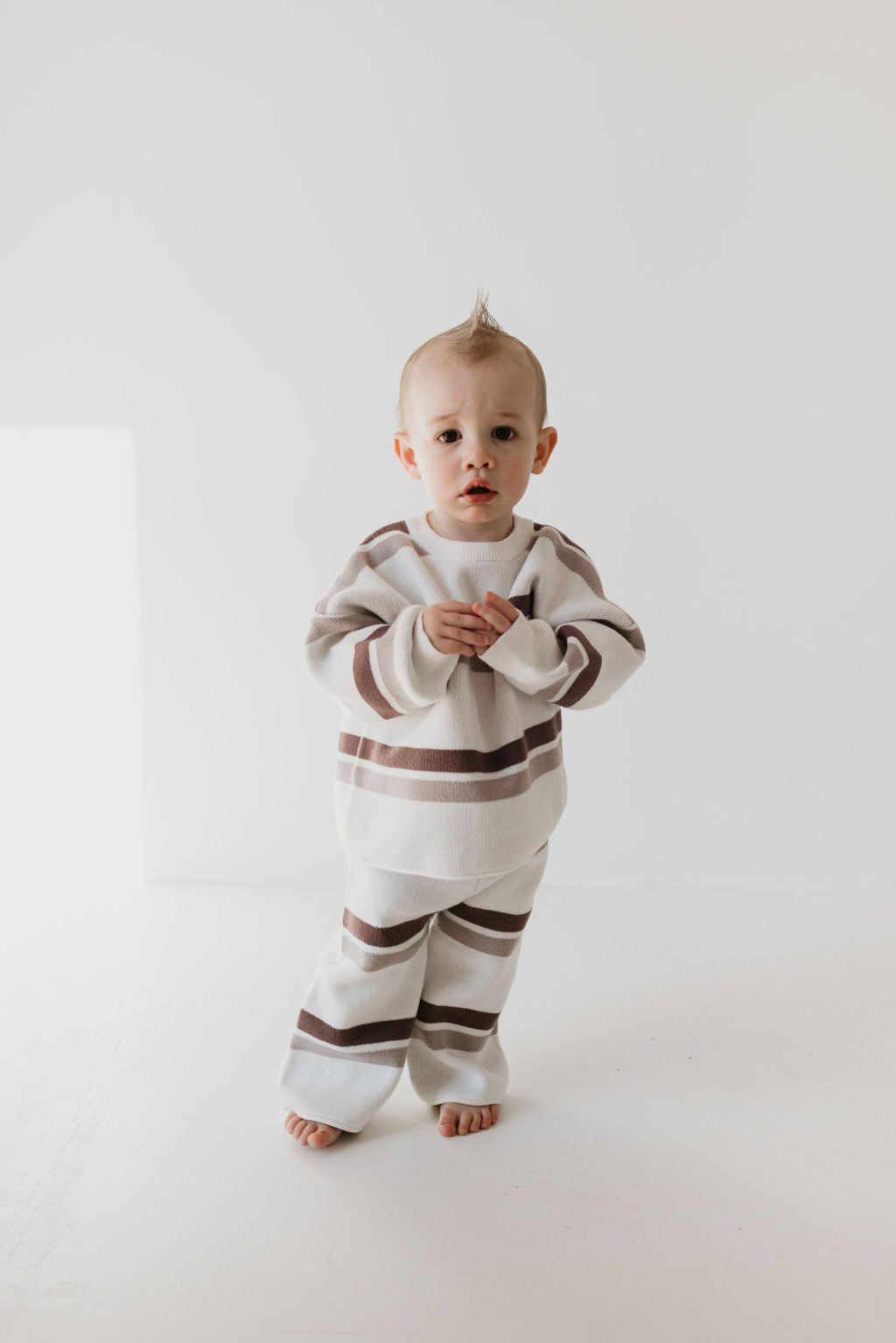 A toddler stands barefoot on a white surface, dressed in the coziest Knit Pant Set | Woodland Stripe by forever french baby. Made from 100% cotton, this outfit is an ideal addition to a fall wardrobe. With a tuft of hair pointing upward, the child gazes directly at the camera with a curious expression.