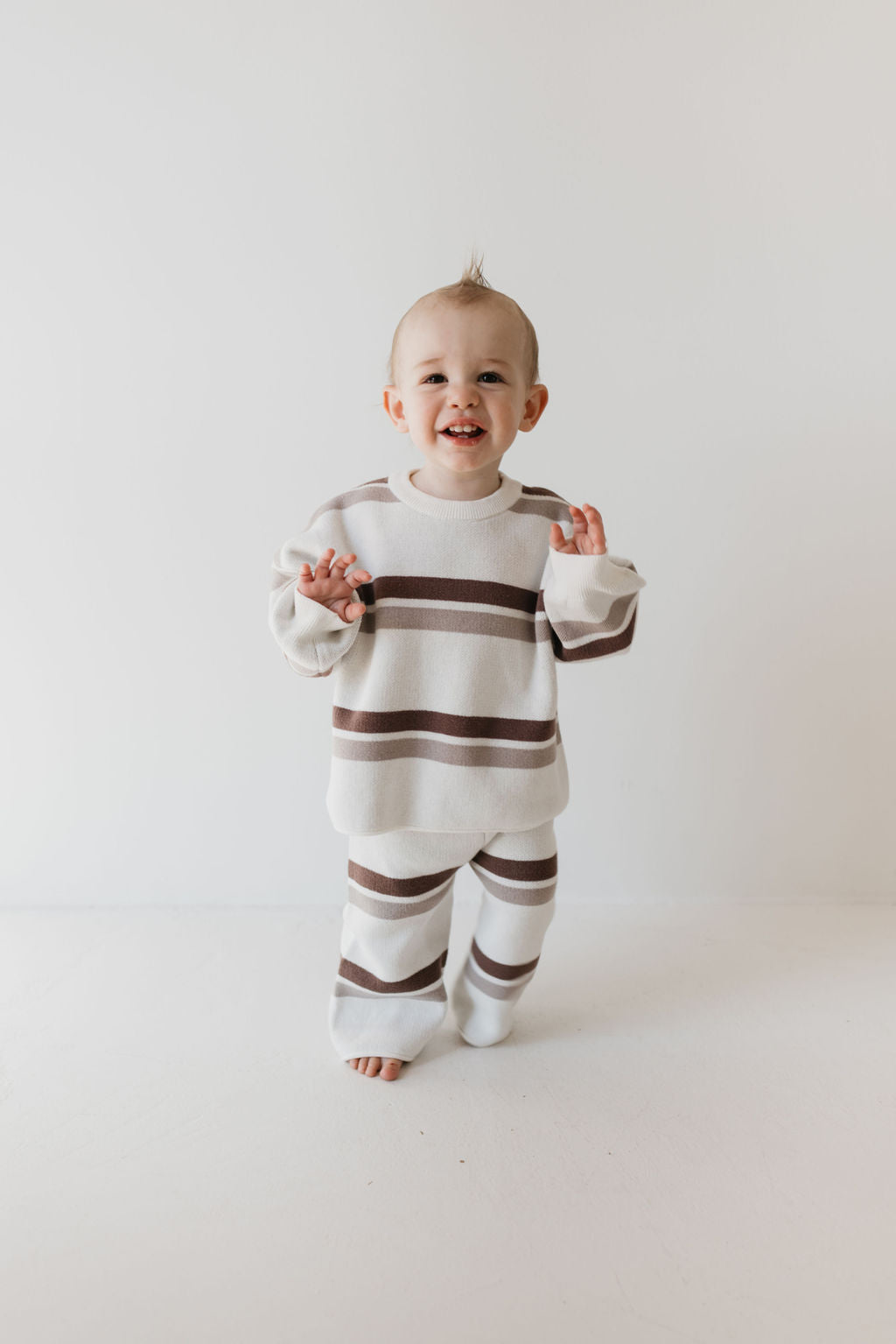 A toddler stands against a plain white background, wearing the charming Knit Pant Set in Woodland Stripe by forever french baby. The child is smiling with hands slightly raised, showcasing the perfect addition to any fall wardrobe, and has short, light hair.