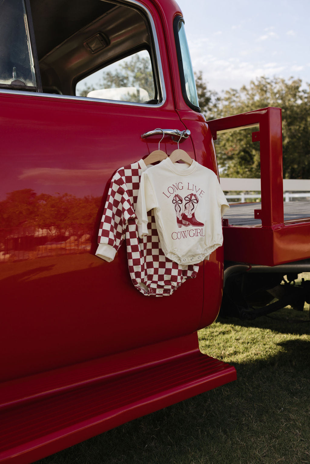 Two baby outfits hang on a red vintage truck's door. One is a sweatshirt romper featuring red and white checks, ideal for cooler months. The other outfit is a cream-colored Long Sleeve Romper from forever french baby with "Long Live Cowgirl" printed alongside a horse and flowers. The truck is parked on grass with trees in the background.