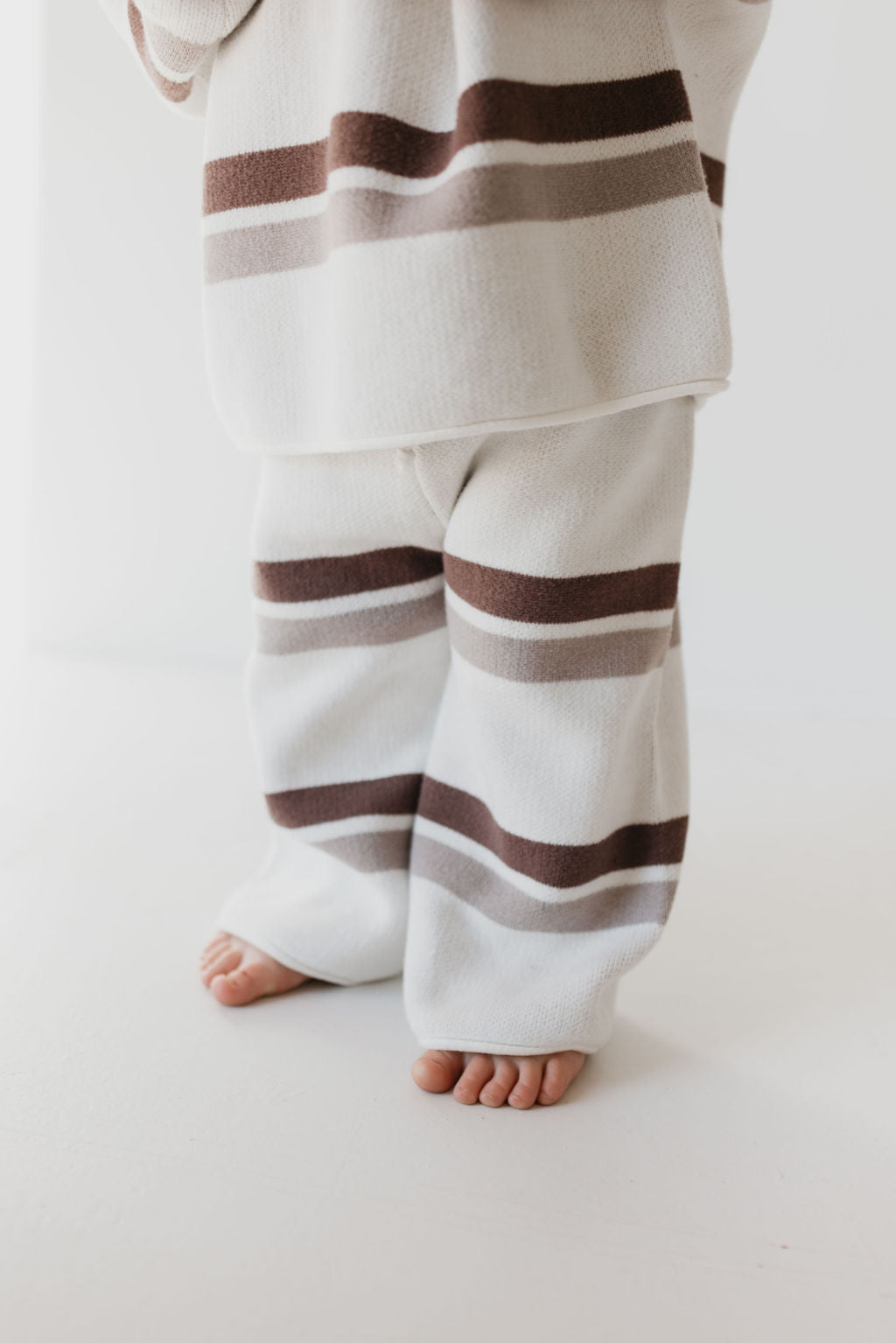 A child in the Knit Pant Set | Woodland Stripe from forever french baby stands barefoot on a light surface, showcasing the cozy and stylish cream-colored knit with brown and gray horizontal stripes—an essential piece for any fall wardrobe.