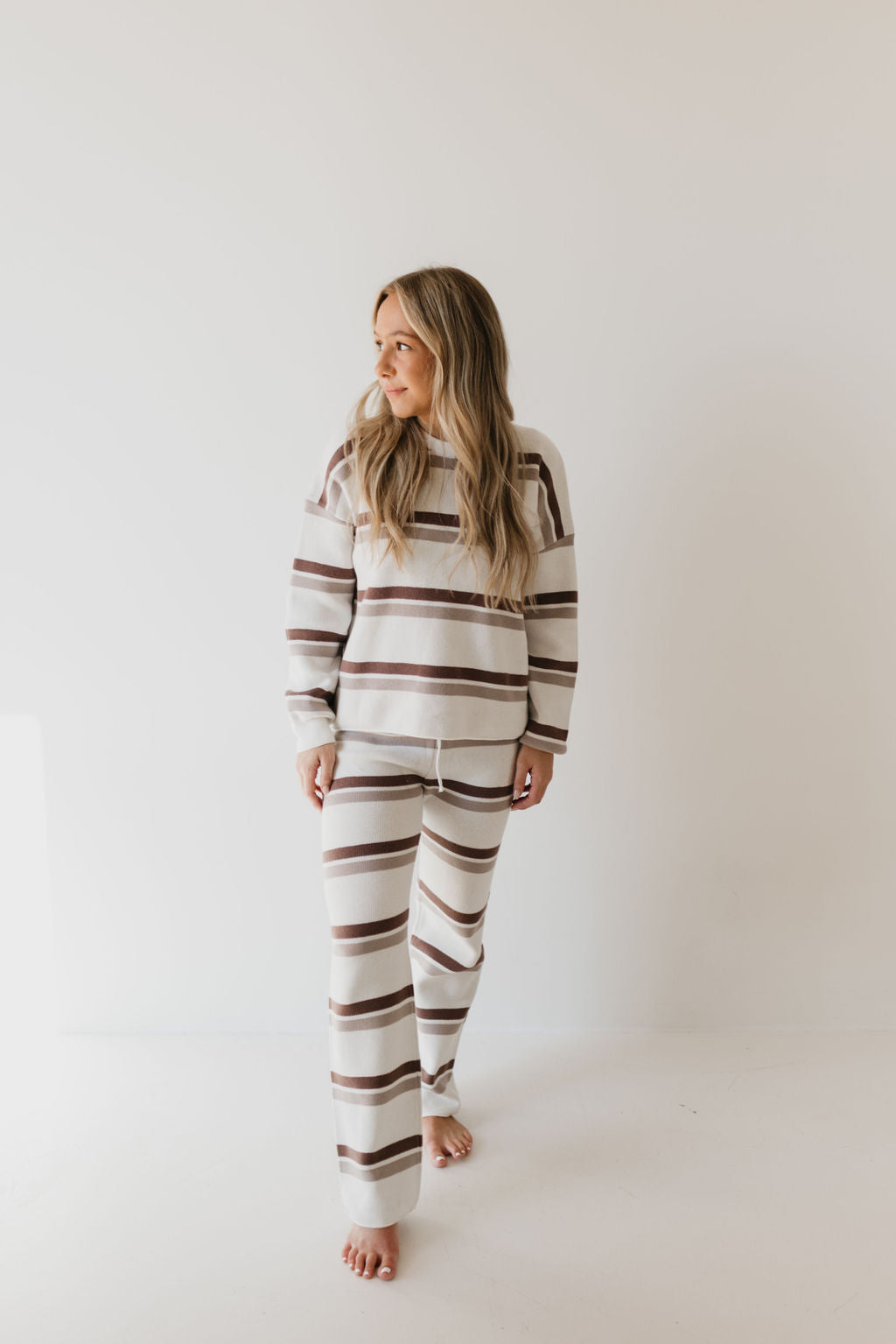 A person with long hair stands barefoot, showcasing the Women's Knit Pant Set in Woodland Stripe by forever french baby, an ideal addition to a fall wardrobe. The set, made from 100% cotton stripes in brown and beige on a cream background, perfectly complements the minimal and light-colored setting. They gaze to the side with a relaxed posture.