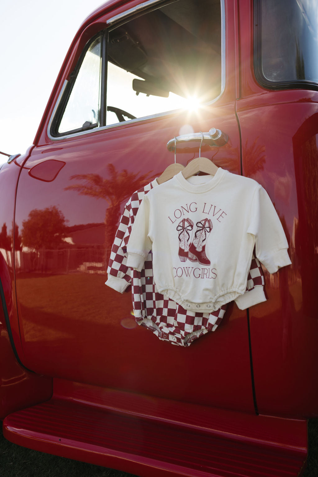 A red vintage truck showcases an adorable outfit from forever french baby: a Long Sleeve Romper with "Long Live Cowgirls" hanging on its door alongside checkered overalls, making it perfect for the cooler months. The sun streaming through the truck's window adds a warm glow to the scene.