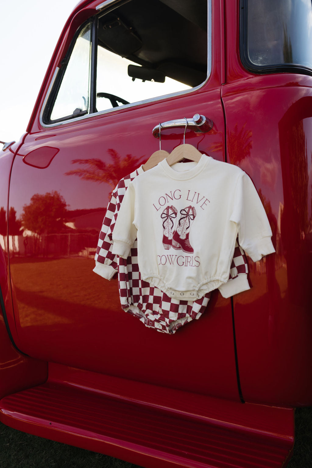 A red vintage truck features a baby outfit hanging on the door, showcasing the Long Sleeve Romper from forever french baby. This sweatshirt romper has a white top with a "Long Live Cowgirls" and cowgirl boots design, complemented by red and white checkered overalls, making it ideal for cooler months.