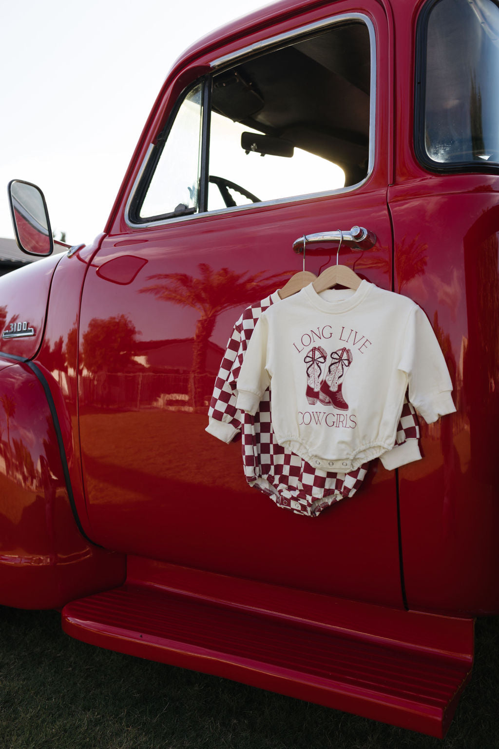 A red vintage truck with a baby outfit hanging on the side mirror highlights a "Long Live Cowgirls" long sleeve romper by forever french baby. This cozy attire is ideal for cooler months, featuring a white top with checkered accents and matching shorts. In the background, trees stand tall under a clear sky.