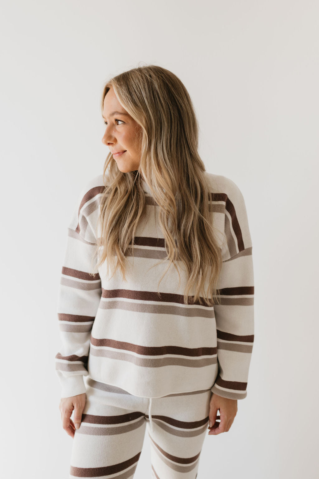 A woman with long blonde hair stands against a plain background, dressed in the Women's Knit Pant Set in Woodland Stripe. Designed by forever french baby, she looks to her left, exuding relaxed elegance—making it a perfect addition to any fall wardrobe.