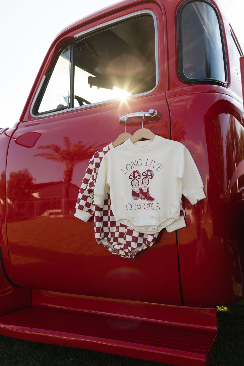 A red vintage truck basks in the sunlight, its window glowing brightly. Draped on the door is a baby outfit featuring the Long Sleeve Romper from forever french baby, with a checkered shirt and a one-piece adorned with "LONG LIVE COWGIRLS" above boot graphics. Ideal for cooler months, it's as charming as the palm trees swaying in the background.