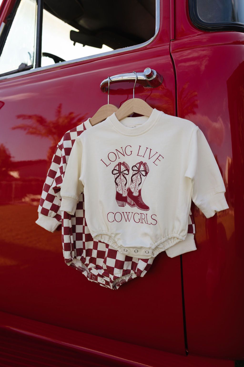 A cream romper from forever french baby, featuring the "Long Live Cowgirls" design with cowboy boots, hangs on a red truck door. Behind it, a red and white checkered shirt can be seen sharing the hanger, ideal for cooler months when combined with a cozy long sleeve romper.