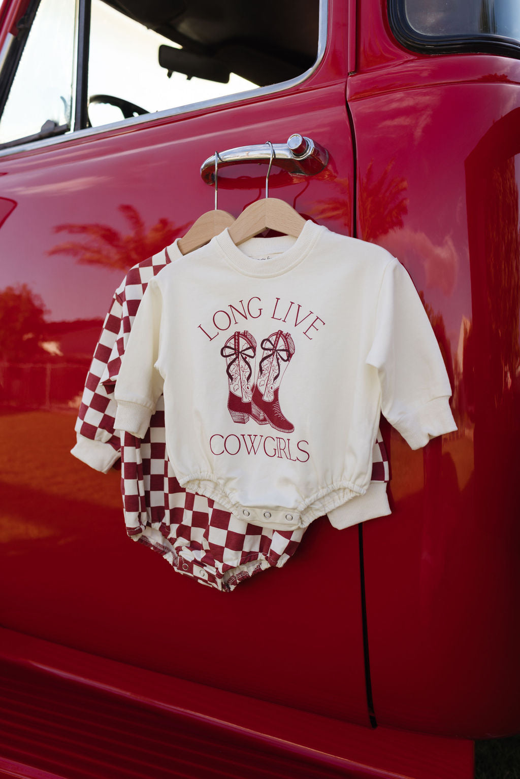 A red vintage truck displays two delightful baby outfits on its door. One of the outfits, a cream-colored long sleeve romper from forever french baby, is adorned with the slogan "Long Live Cowgirls" alongside a cowboy boots print—perfect for cooler months. The other outfit features a timeless red and white checkered design.