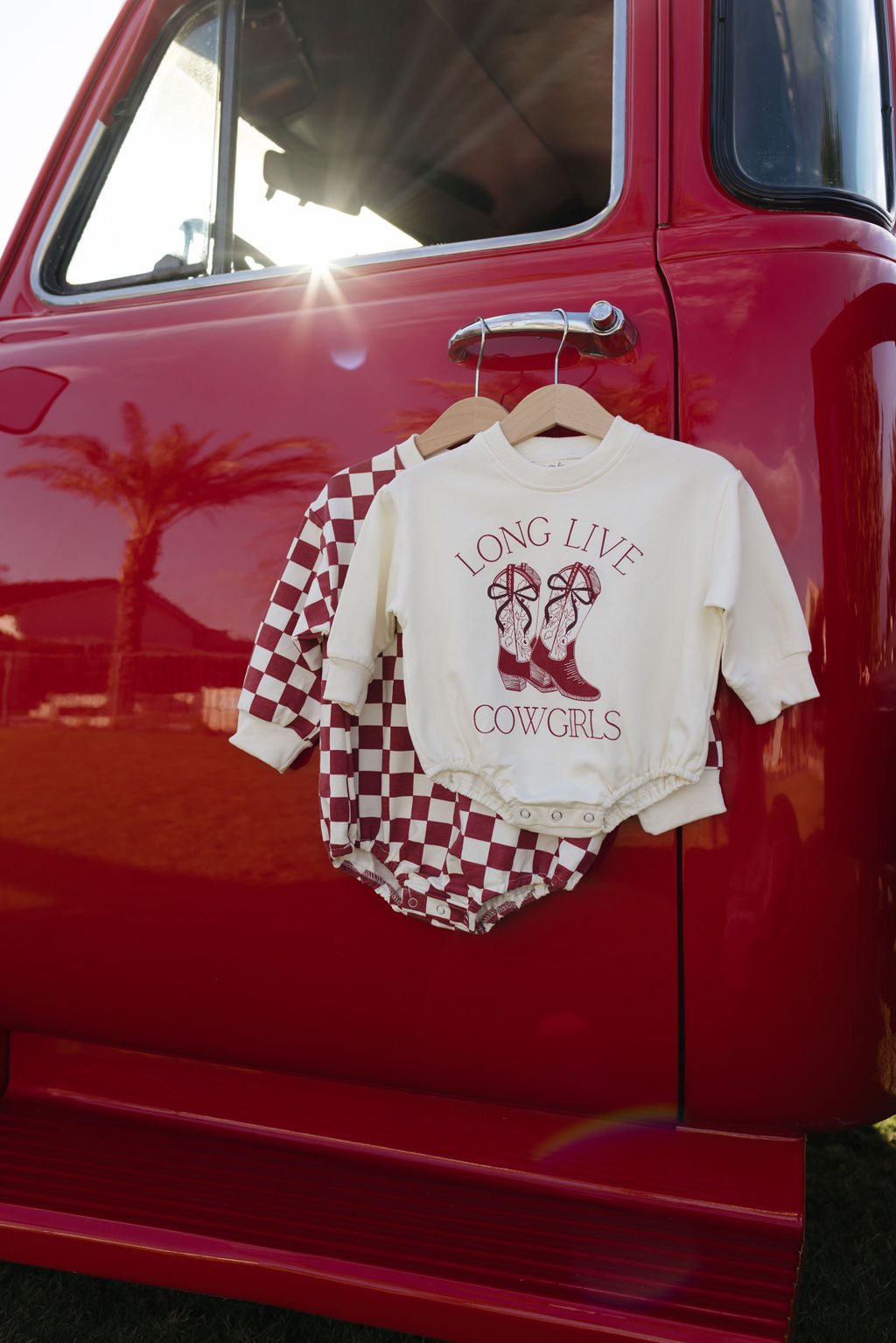 A red vintage truck showcases a checkered baby outfit alongside a sweatshirt romper, with a cream "Long Live Cowgirls" long sleeve romper from forever french baby hanging on its window. Sunlight pours through, signaling the approach of cooler months.