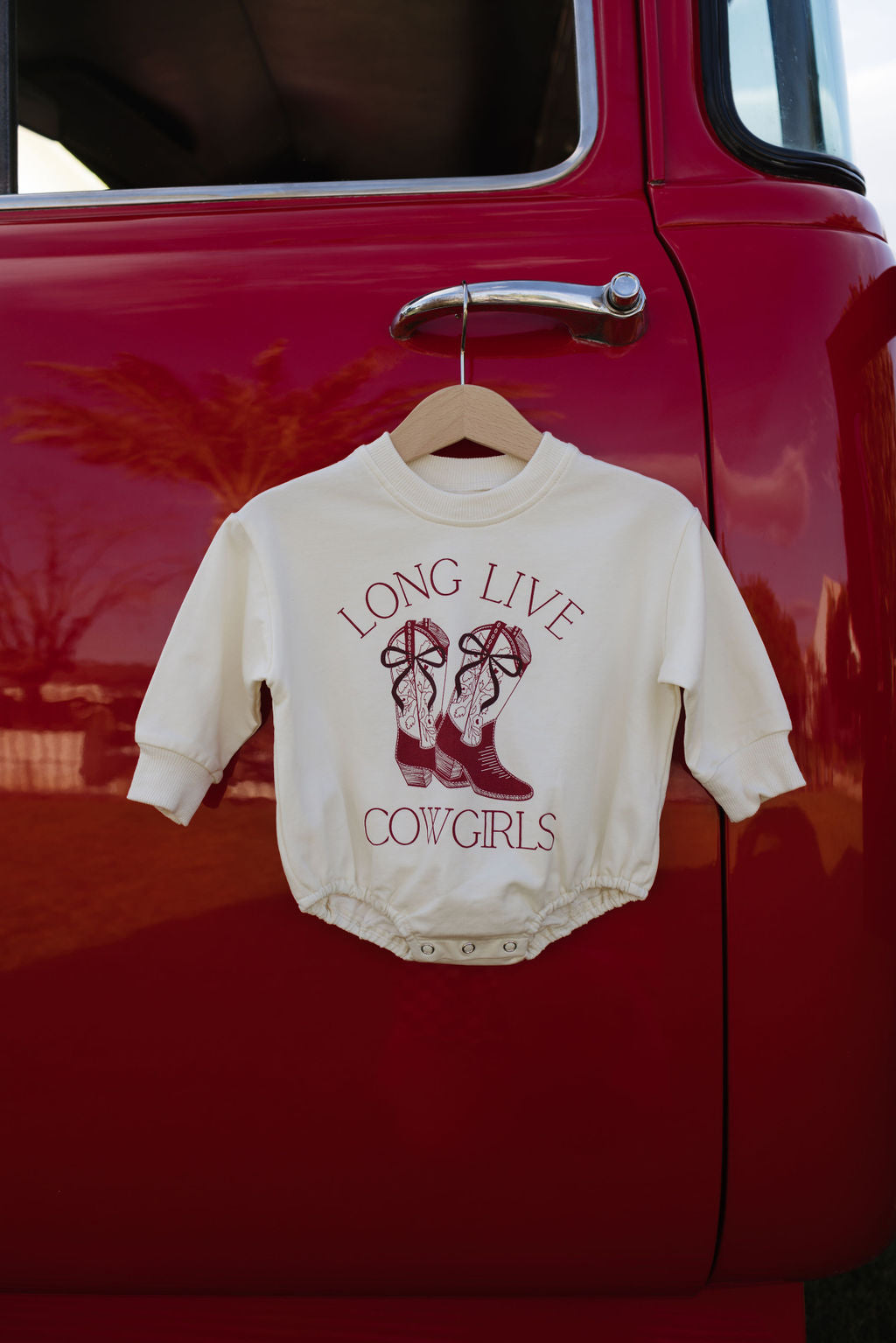 A cream-colored child's long sleeve romper with the "Long Live Cowgirls" and cowboy boots design by forever french baby is draped on a wooden hanger from the side mirror of a red vintage truck, ideal for cooler months.