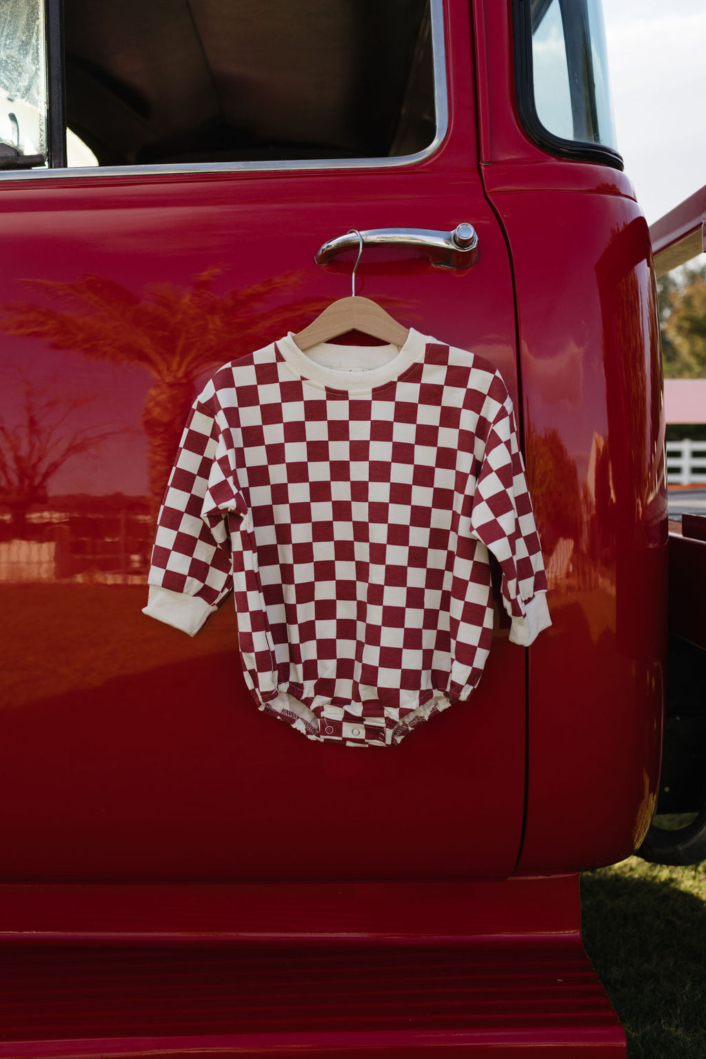 A Long Sleeve Romper from lolo webb, known as "the Quinn," featuring a red and white checkered design that is ideal for newborns to 24 months during cooler months, is displayed on a hanger against the door of a red vehicle, with trees reflected in the vehicle's window.