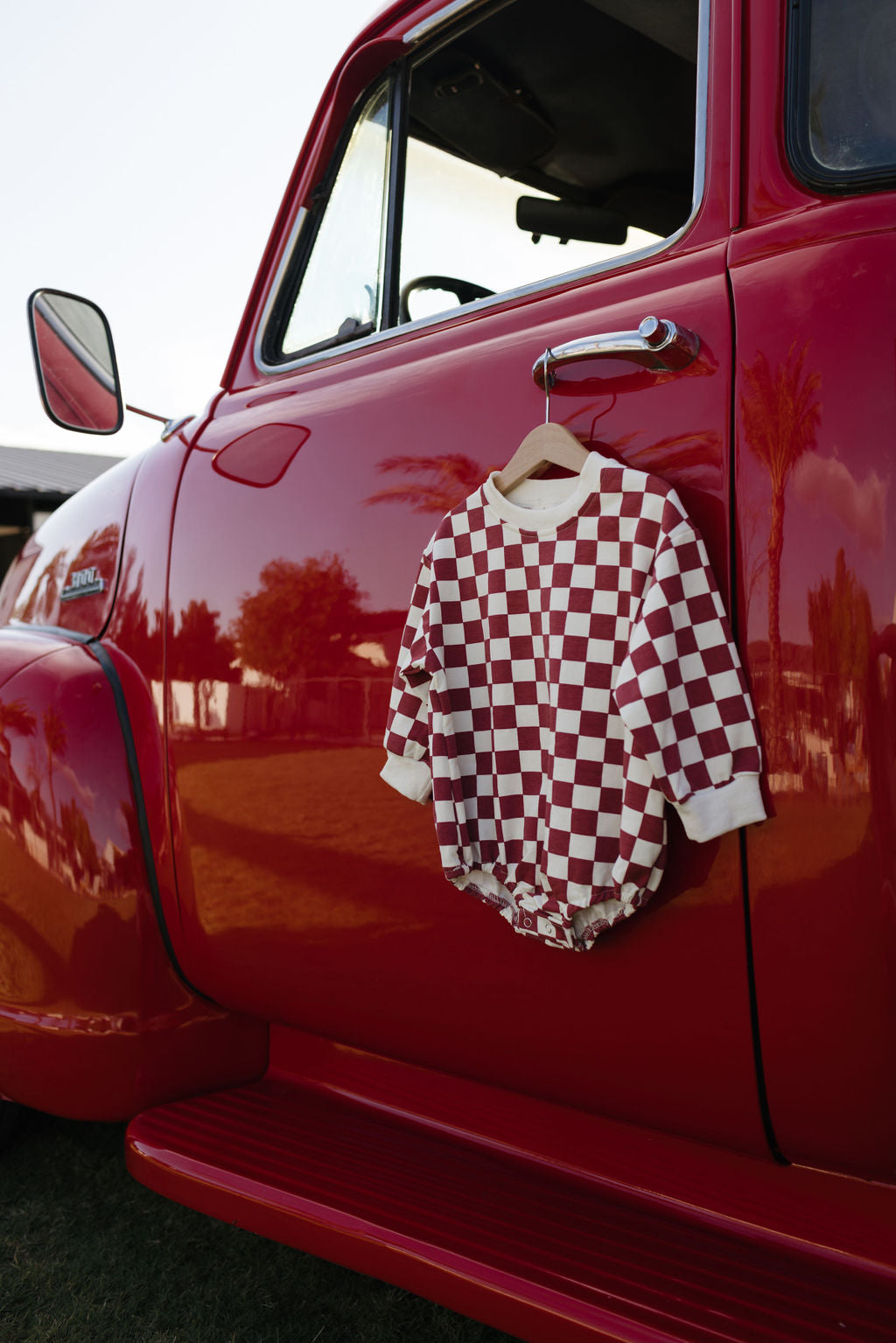 A red and white checkered Long Sleeve Romper from lolo webb's Quinn collection, ideal for newborns to 24 months, hangs on the side mirror of a vintage red truck. The truck is parked in a sunlit outdoor setting with trees in the background, creating a cozy atmosphere for cooler months.