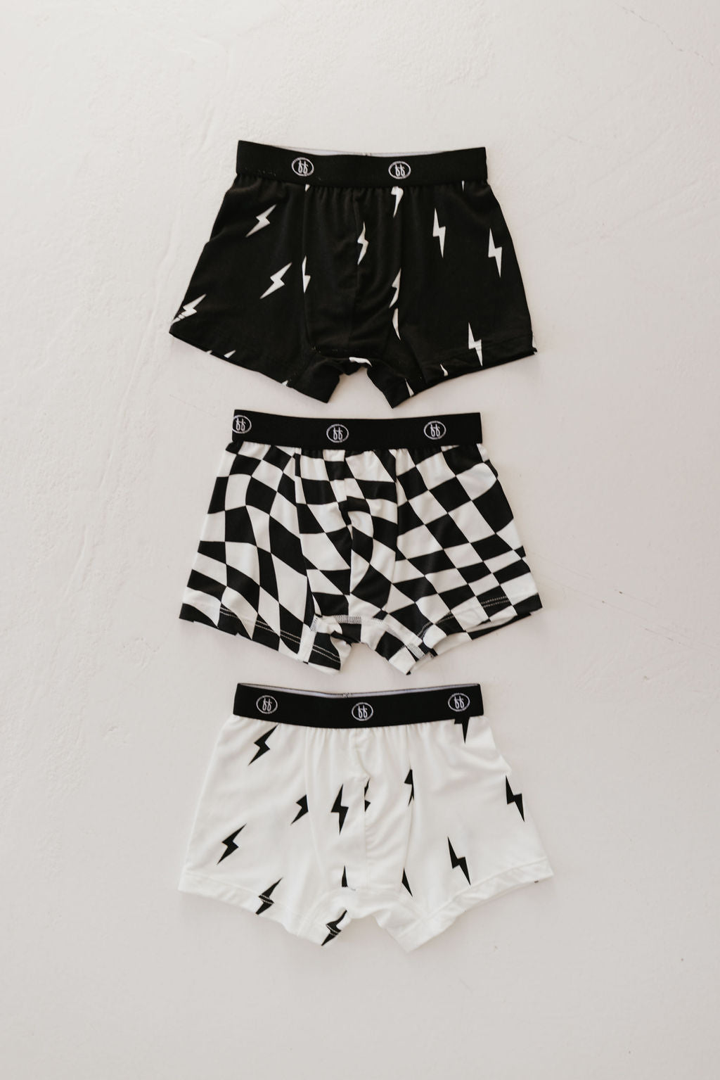 Three pairs of forever french baby's Bamboo Boxers from the "Black and White Lightning Bolt & Wavy Checkerboard" 3-pack are displayed on a light surface. The top pair is black with white lightning bolts, the middle pair showcases a black and white checkered pattern, and the bottom pair is white adorned with bold black lightning bolts.