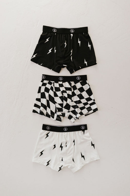 Three pairs of forever french baby's Bamboo Boxers from the "Black and White Lightning Bolt & Wavy Checkerboard" 3-pack are displayed on a light surface. The top pair is black with white lightning bolts, the middle pair showcases a black and white checkered pattern, and the bottom pair is white adorned with bold black lightning bolts.