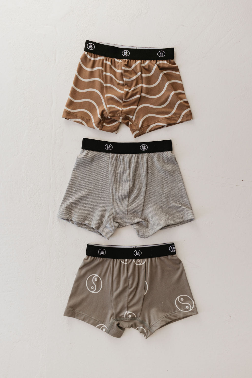 Three pairs of men's breathable boxer briefs from the Bamboo Boxers (3 Pack) by forever french baby are displayed on a light surface. One pair showcases a wavy brown pattern, another is plain gray, and the third features a yin-yang design. Made from hypo-allergenic bamboo fabric, all boxer briefs come with black waistbands adorned with a circular logo.