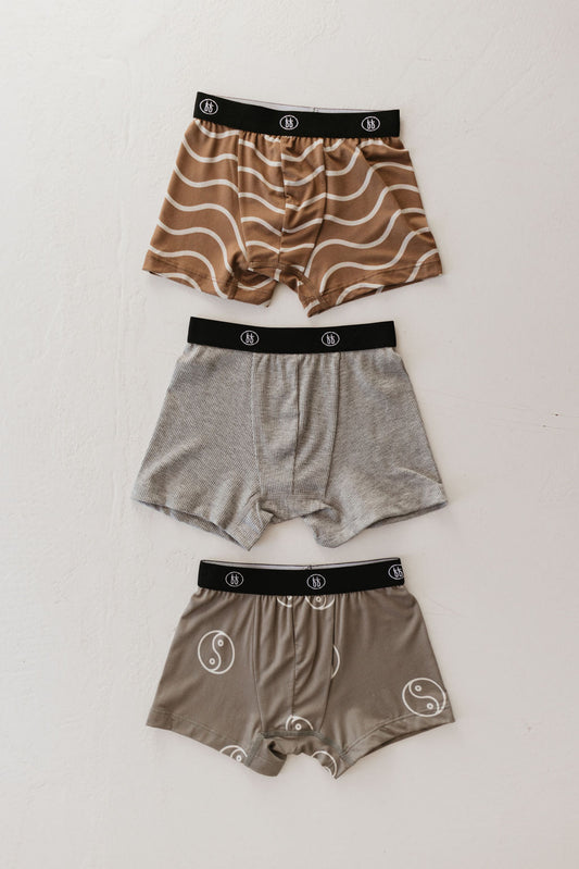 Three pairs of men's breathable boxer briefs from the Bamboo Boxers (3 Pack) by forever french baby are displayed on a light surface. One pair showcases a wavy brown pattern, another is plain gray, and the third features a yin-yang design. Made from hypo-allergenic bamboo fabric, all boxer briefs come with black waistbands adorned with a circular logo.