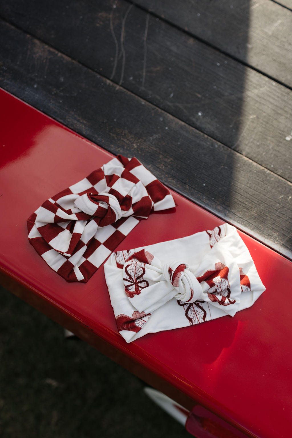 Two knotted hairbands rest on a red wooden surface. One, the Bamboo Head Wrap by lolo webb called "the Quinn," displays a red and white checkered pattern, while the other shows off a white base with vibrant red floral designs. The scene is set against a contrasting dark wooden tabletop in the background.