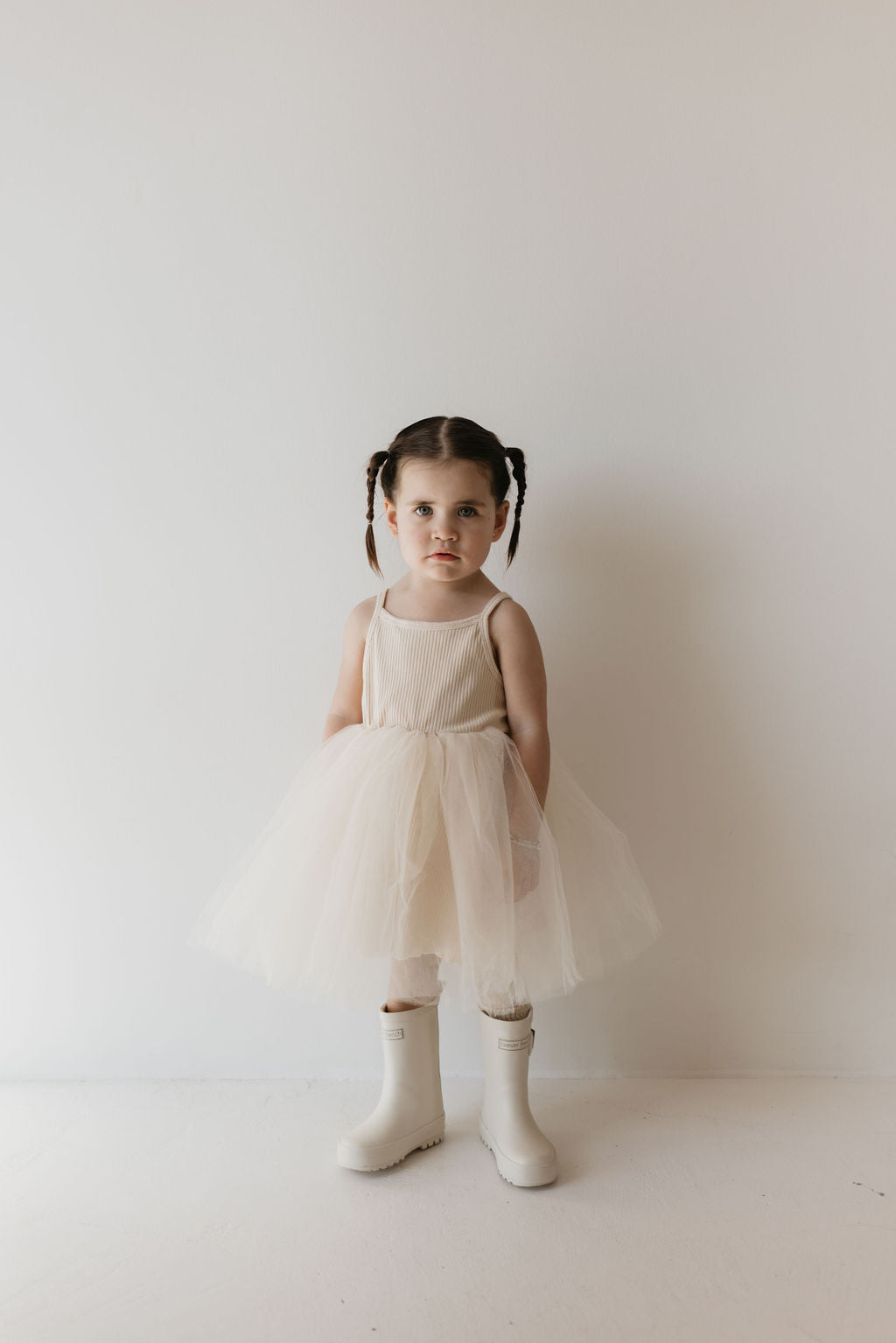 A young child stands against a plain white background, wearing the Sugar ff Tutu Dress from forever french baby, paired with white leggings and signature ff rain boots. The child's hair is styled in two braids, showcasing a classic kid's wardrobe staple.