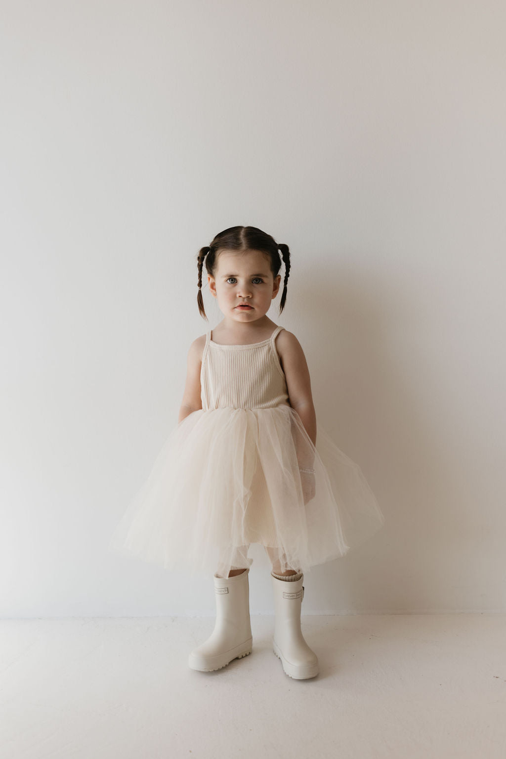 A young child with pigtails is dressed in the forever french baby ff Tutu Dress | Sugar, a kid's wardrobe staple, paired with signature FF rain boots. They stand against a plain, white background, looking directly at the camera.