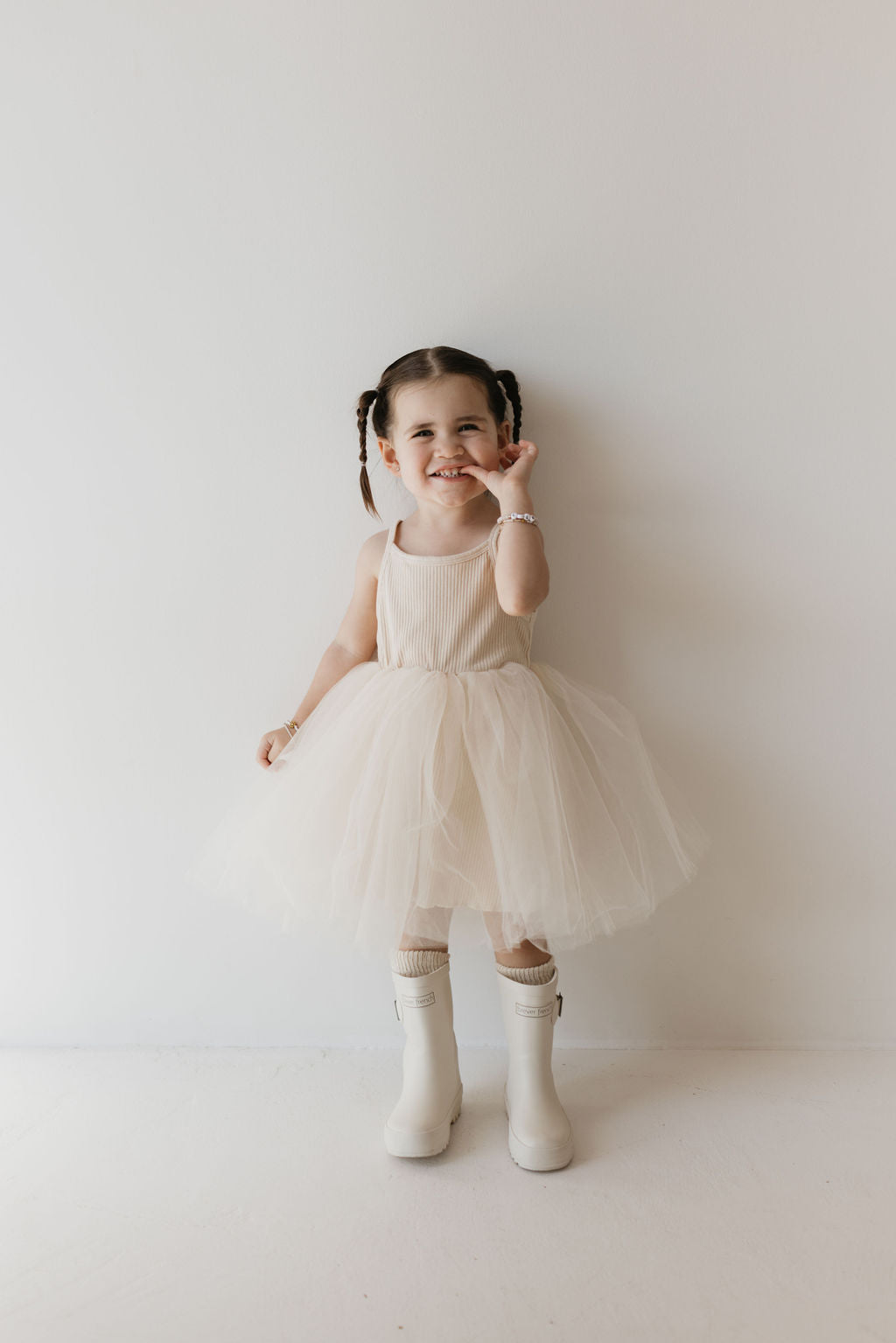 A young child with pigtails stands against a white wall, wearing the elegant ff Tutu Dress in the Sugar color by forever french baby and their signature ff rain boots. Smiling brightly with one hand close to their face, the child showcases this kid's wardrobe staple with charm.