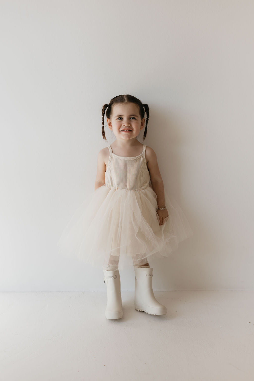 A young girl with braided hair smiles, wearing the ff Tutu Dress in Sugar by forever french baby and signature ff rain boots. She stands against a plain white background, showcasing effortless charm—a true kid's wardrobe staple.