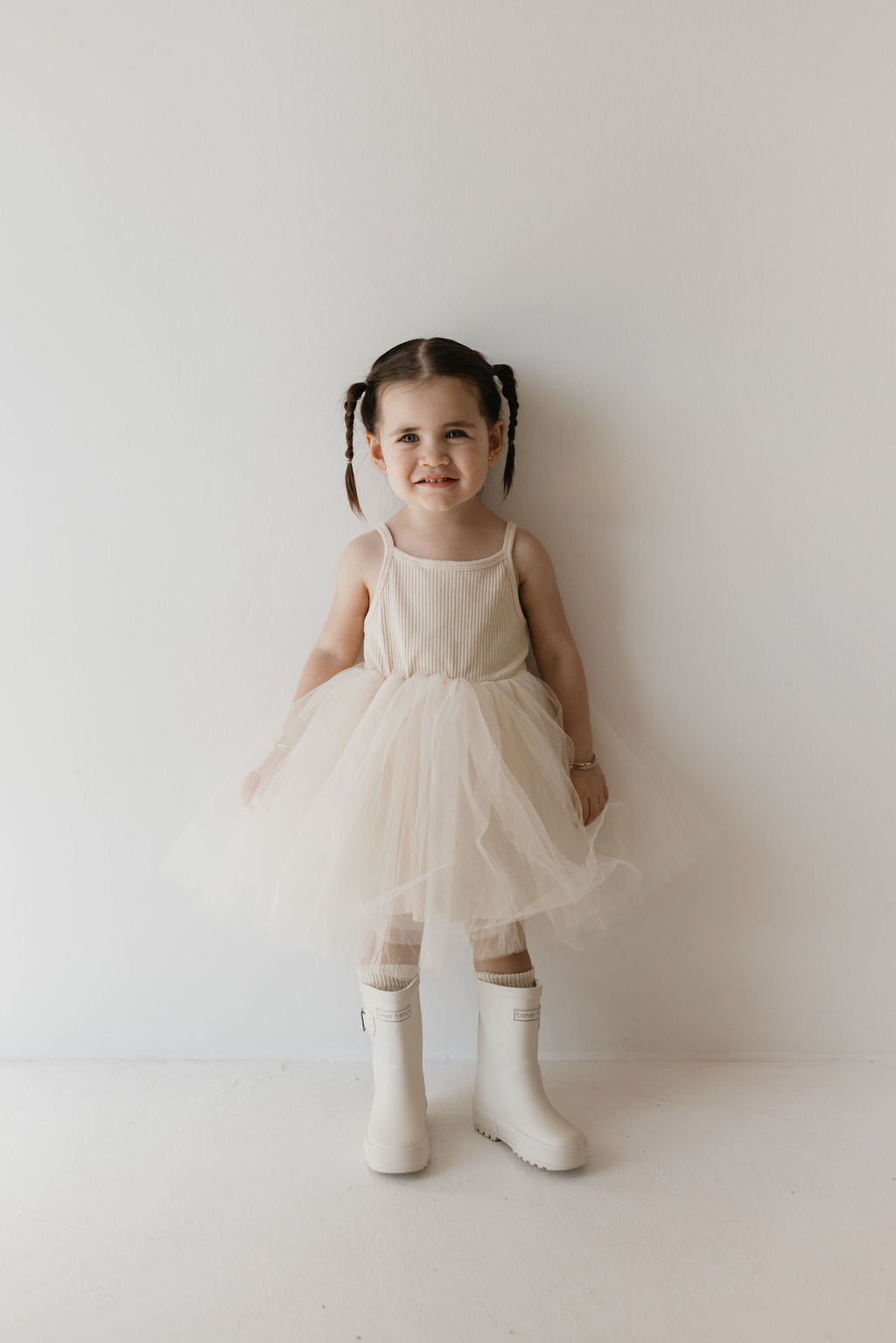 A young child stands against a plain white background, smiling. She is wearing the ff Tutu Dress in Sugar by forever french baby, a staple in any kid's wardrobe, paired with signature FF rain boots. Her hair is styled in pigtails, and she exudes a playful demeanor.