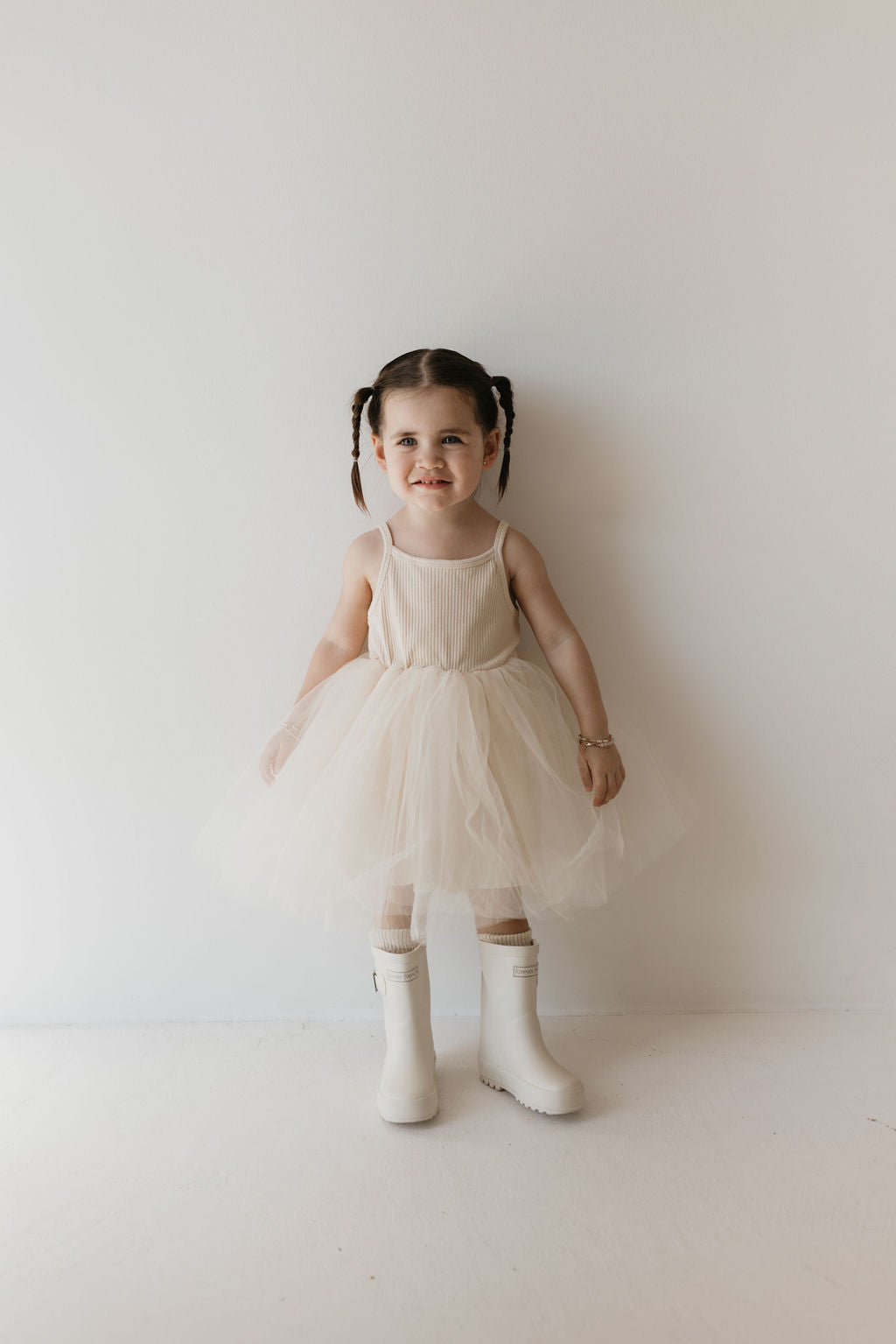 A young child with dark braided hair stands against a plain white background, donning a cream-colored sleeveless ff Tutu Dress | Sugar from forever french baby, paired with matching signature ff rain boots. With a slight smile, she looks directly at the camera, capturing an iconic kid's wardrobe staple moment.