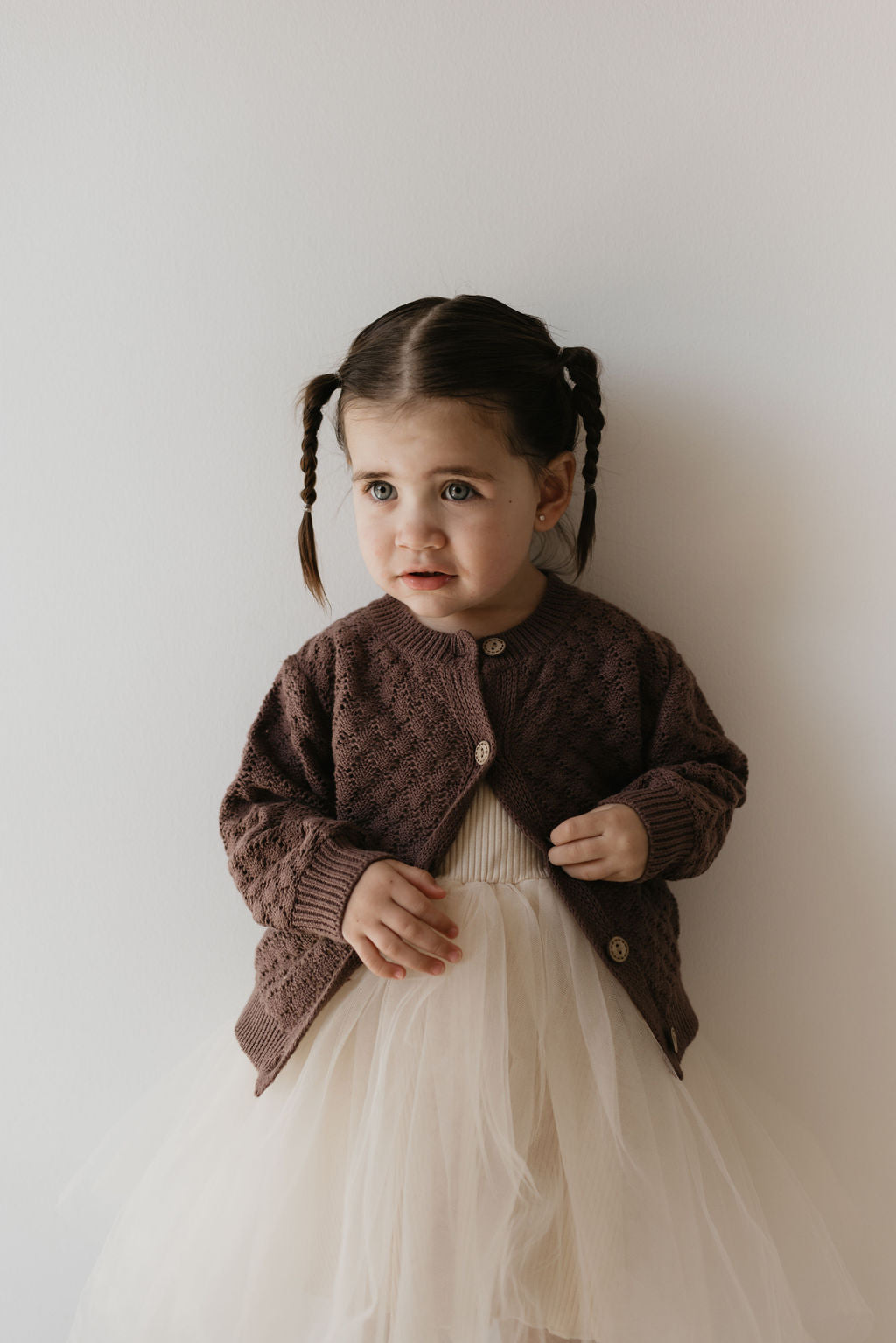 In front of a simple backdrop, a young child with braided hair wears the Knit Cardigan in Clove from Forever French Baby, crafted from breathable cotton. Paired with a cream-colored tulle dress, they gaze slightly to the side with a gentle expression, highlighting versatile styling.