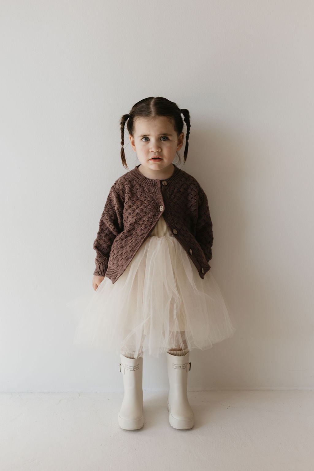 A young child with braided hair stands against a neutral wall, wearing the Clove Knit Cardigan from forever french baby, made of breathable cotton. The ensemble is completed with a cream-colored tulle skirt and white boots. They look directly at the camera with a neutral expression, showcasing versatile styling.