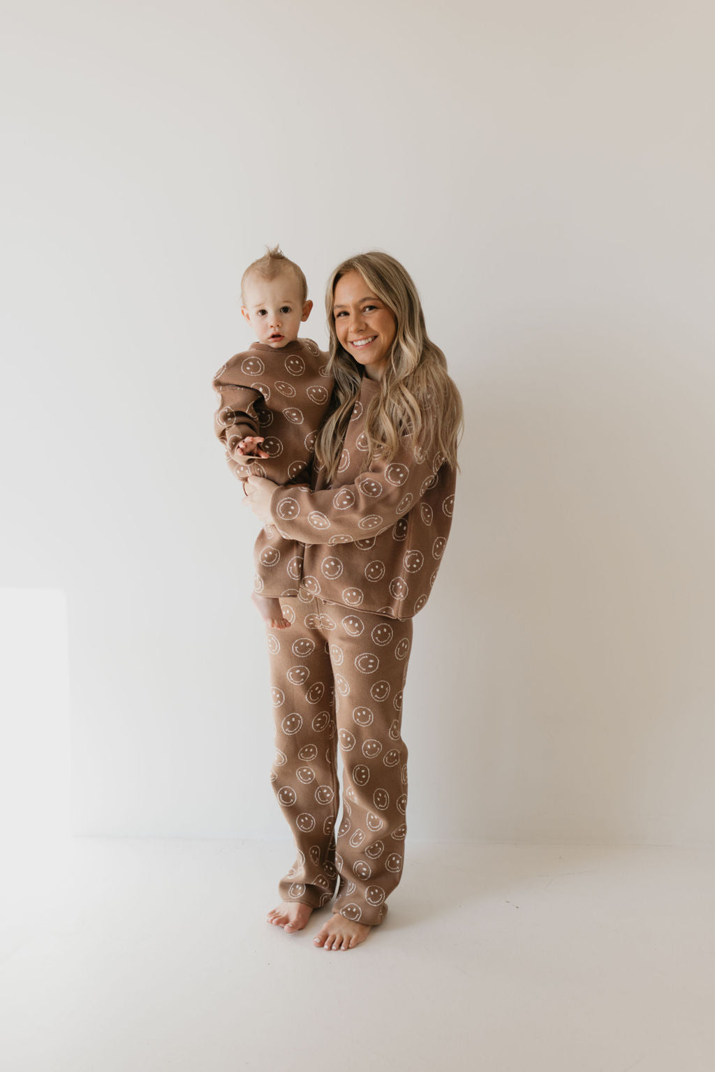 In a minimalist room with a white background, a woman and her baby are standing together, both dressed in matching outfits from the "Women's Knit Pant Set | Tan Just Smile" collection by forever french baby. The woman is smiling as she holds the baby, who is looking at the camera. Their outfits feature playful smiley face patterns and are made from 100% cotton.