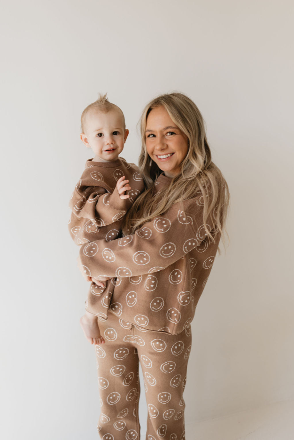 A woman holding a toddler is dressed in matching outfits from forever french baby's "Women's Knit Pant Set | Tan Just Smile," against a plain white background. The oversized top and the knit set, both made from 100% cotton, enhance their coordinated and comfortable style as they smile at the camera.