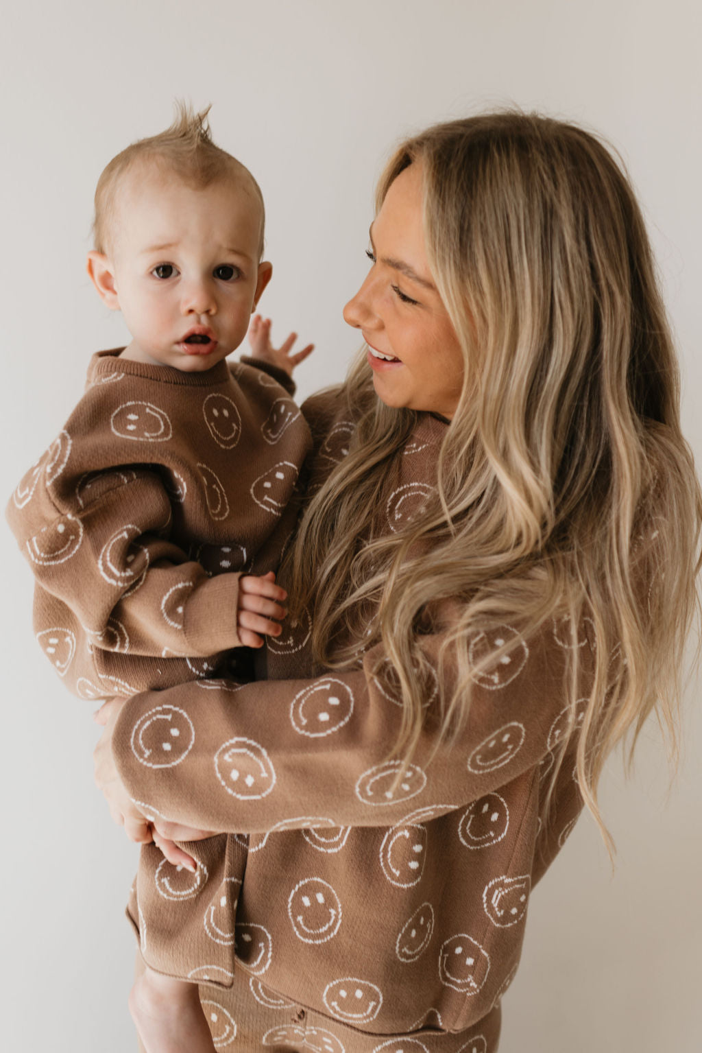 A woman with long blonde hair cradles a baby, both dressed in the Women's Knit Pant Set | Tan Just Smile by forever french baby, featuring matching brown sweaters adorned with cheerful white smiley faces. The woman gazes lovingly at the baby, whose tiny tuft of hair stands up charmingly against the plain, light-colored wall—making it a perfect addition to any fall wardrobe.