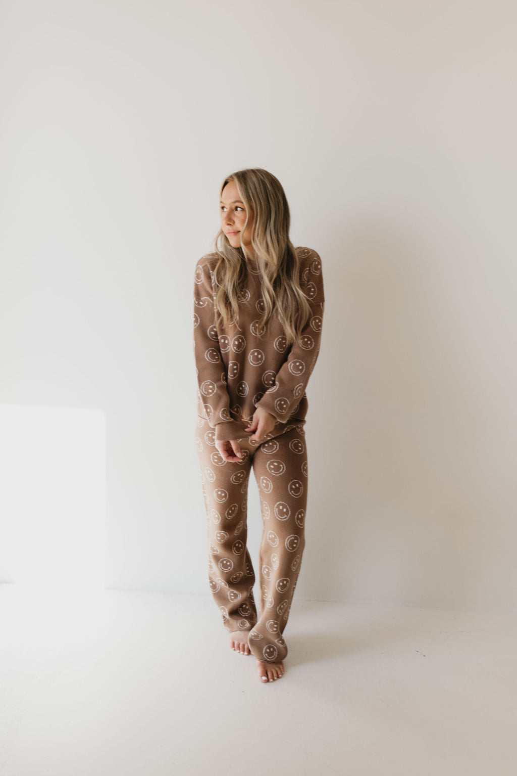 A person with long hair stands barefoot against a white wall, wearing the Women's Knit Pant Set in Tan Just Smile by forever french baby. This matching set features circular patterns and is made from 100% cotton, making it an ideal addition to your fall wardrobe. They maintain a relaxed posture and gaze to their left, with soft lighting creating an inviting atmosphere.