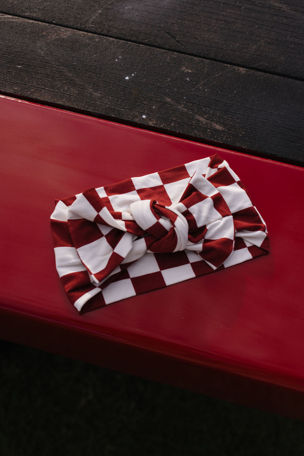 Laid on a red surface, the Bamboo Head Wrap, known as "the Quinn" by lolo webb, features a charming red and white checkered pattern with a knotted design, reminiscent of an adorable baby head wrap. The dark wooden texture in the background enhances the vibrant appearance of this stylish accessory.