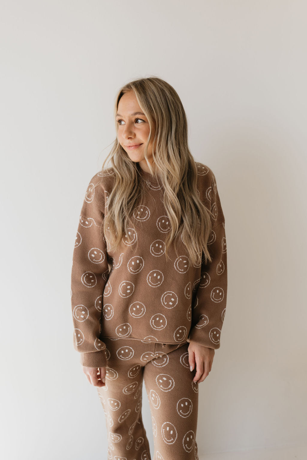 A woman wearing the Women's Knit Pant Set in Tan Just Smile from forever french baby features a smiley face pattern. She gazes to the side with a neutral expression, and her long, wavy blonde hair perfectly complements the oversized top. The background is plain and light-colored.