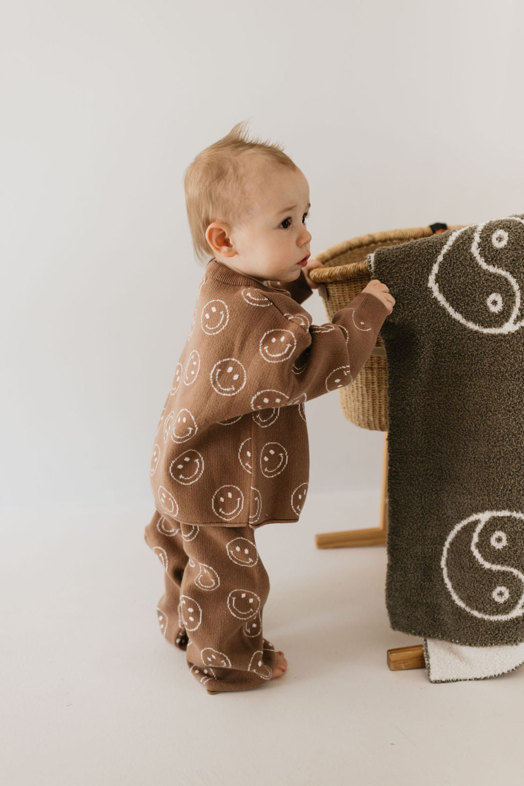 A baby stands and holds onto a basket, dressed in the Knit Pant Set | Tan Just Smile by forever french baby. A cozy brown outfit adorned with white smiley face patterns keeps the little one warm. Draped over the basket is a dark green blanket featuring white yin-yang symbols, enhancing the charm of this delightful fall wardrobe scene against a plain, light-colored background.