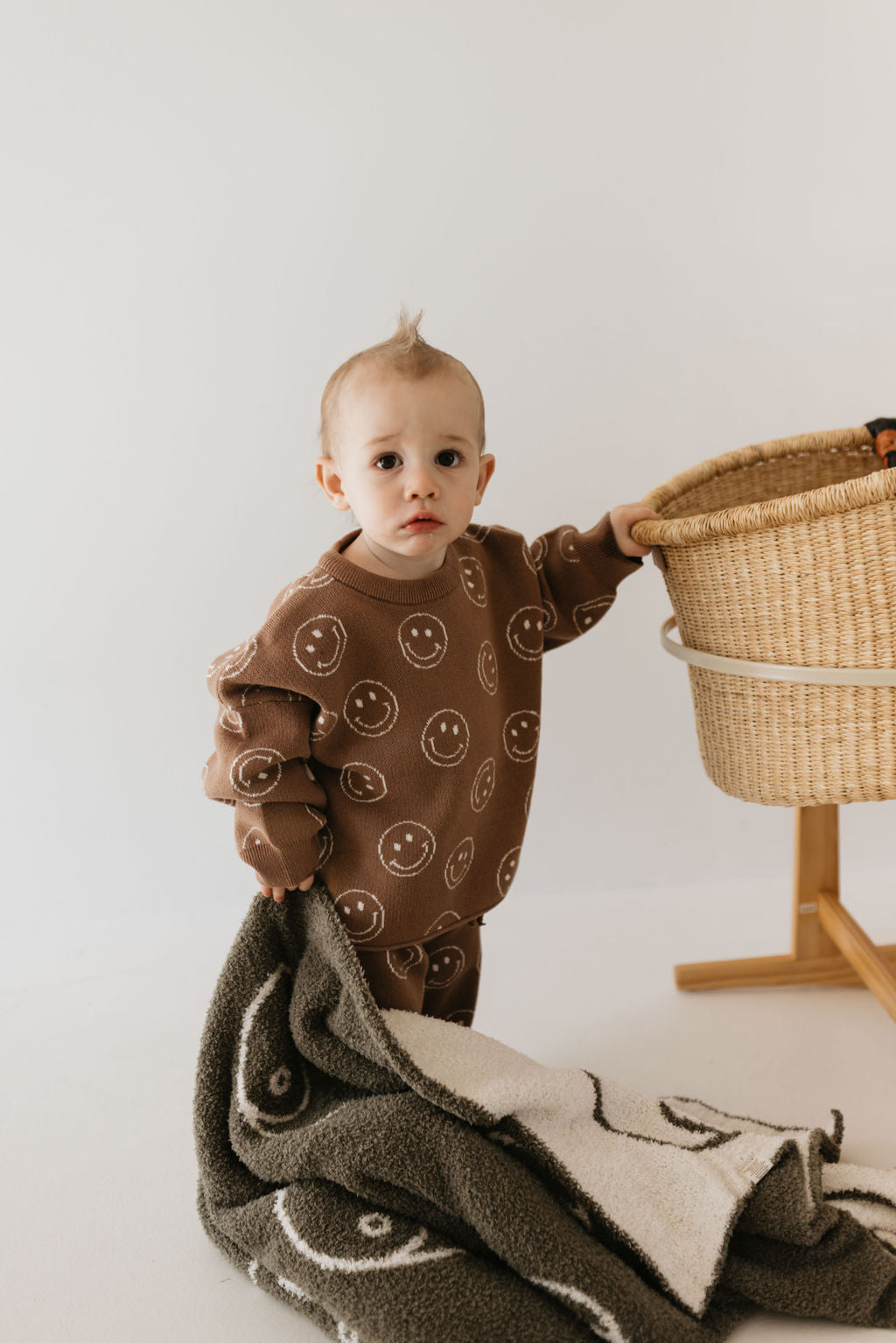 A baby in a brown outfit with smiley faces stands holding a woven basket. In the other hand, the baby clutches a Forever French Baby Plush Blanket in Sage Yin Yang design. The background is plain and light-colored, creating a serene and minimalist setting.