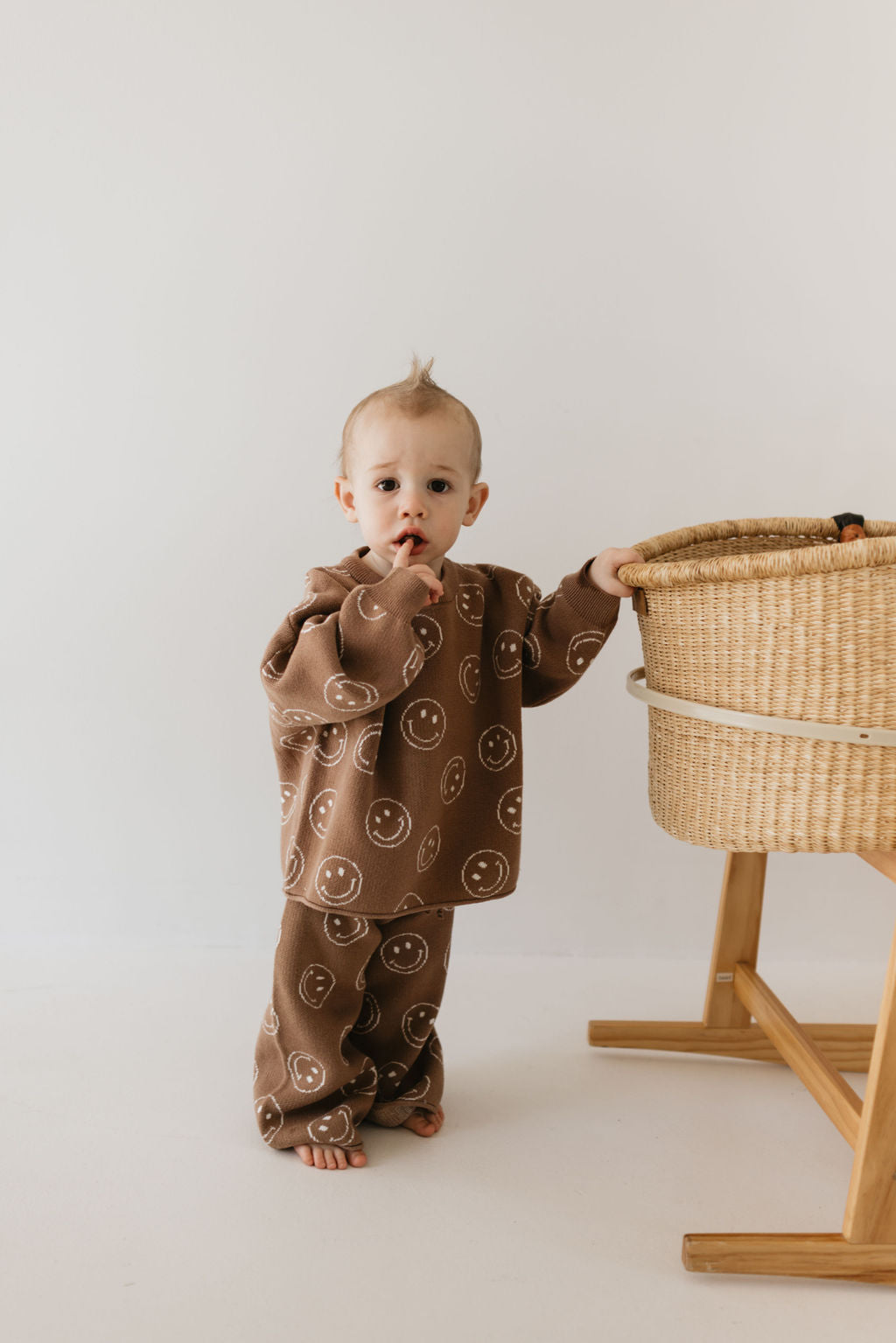 A baby stands next to a woven bassinet on a stand, dressed in the cozy Knit Pant Set | Tan Just Smile from forever french baby, featuring adorable smiley faces. The curious expression and the minimalist backdrop of a plain white wall make this scene delightful for any fall wardrobe inspiration.