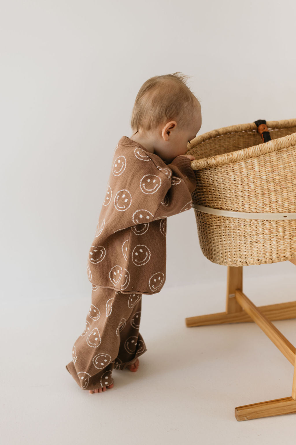 A baby dressed in the Knit Pant Set | Tan Just Smile from forever french baby, featuring a charming pattern of smiley faces, leans against a woven basket bassinet on a wooden stand. This outfit is perfect for the fall wardrobe and made from 100% cotton. The minimalist appeal is enhanced by the plain white wall in the background.