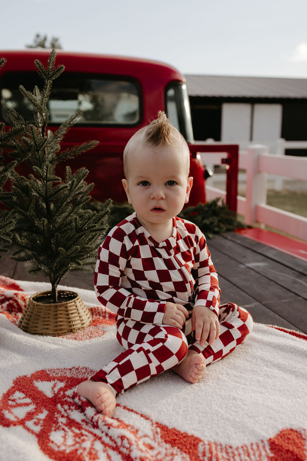 A baby, dressed in "Bamboo Zip Pajamas | the Quinn" by lolo webb, sits on a blanket with a red and white checkered pattern. On a wooden deck near a small pine tree and a red vehicle, the scene is both charming and serene.