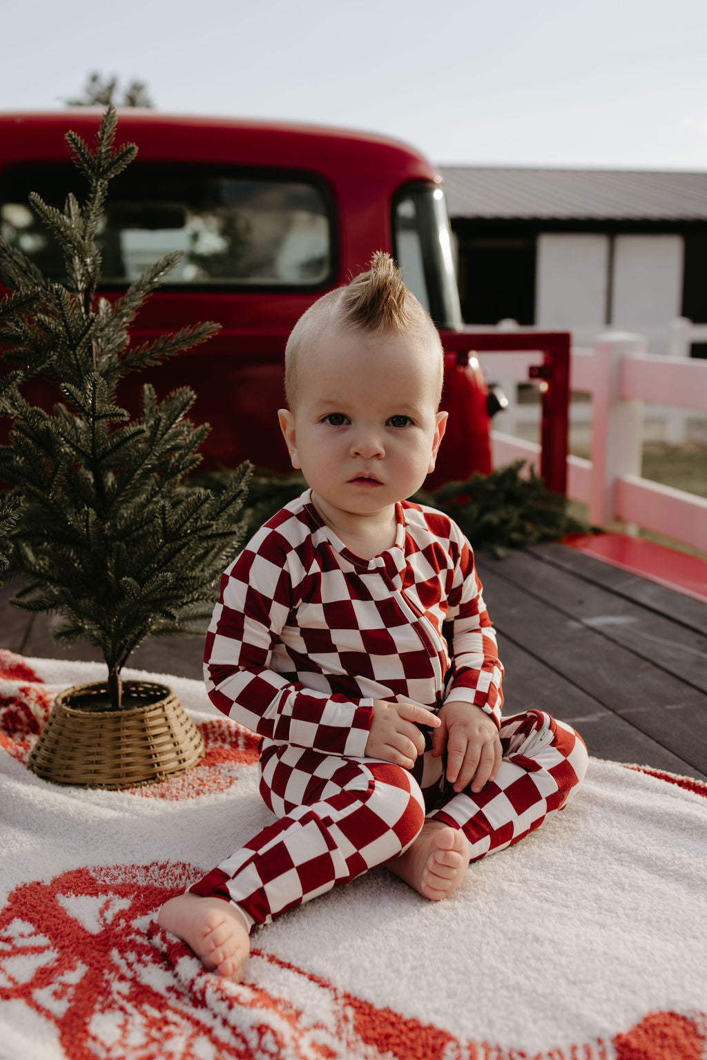 A baby wearing the Quinn bamboo zip pajamas by lolo webb sits on a white and red blanket outdoors. There's a small potted evergreen tree beside them and a red vintage truck in the background, enhancing the festive and outdoorsy scene.