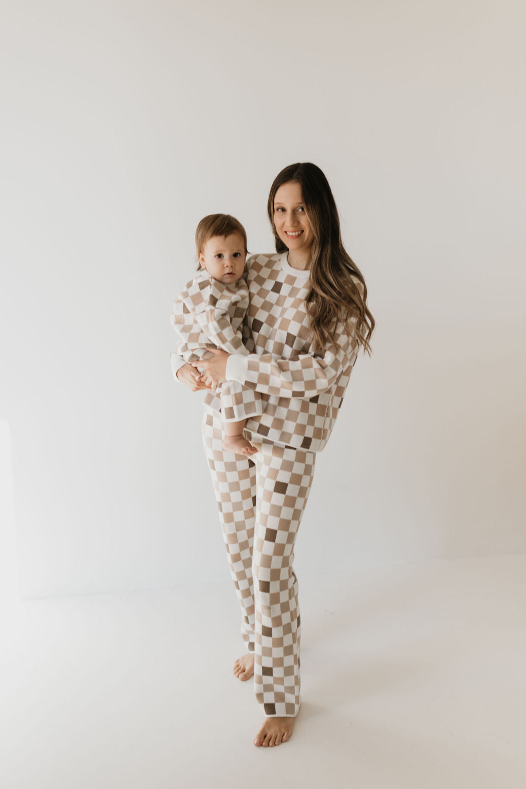 A woman dressed in a Women's Knit Pant Set from the "Into the Woods Checkerboard" collection by Forever French Baby holds her baby, both adorned in matching beige and white checkered pajamas. They stand barefoot on a white background, with the woman smiling at the camera as the baby gazes curiously ahead.