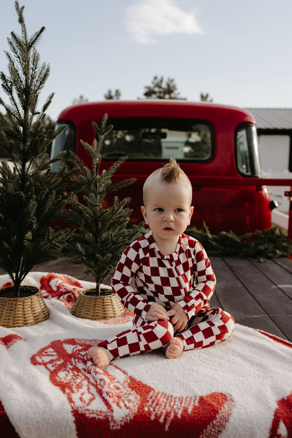 A baby wearing the breathable lolo webb Bamboo Zip Pajamas in a red and white checkered pattern sits on a festive blanket next to small Christmas trees. In the background, a vintage red truck adds charm to the outdoor scene under a clear sky.